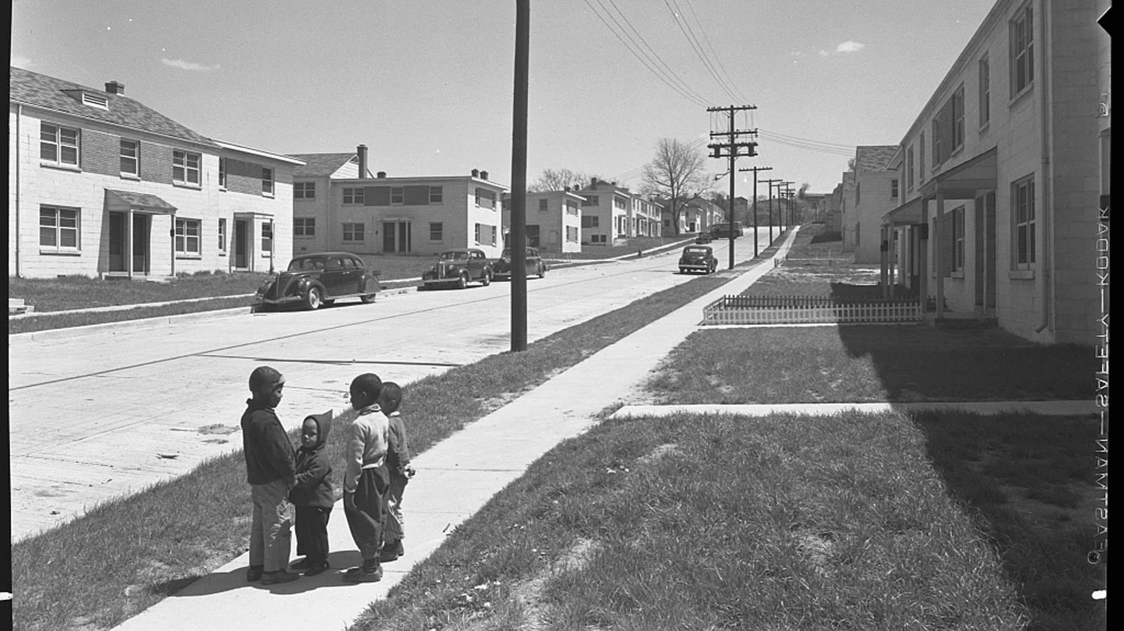 Four African American children stand on a sidewalk in a housing development.