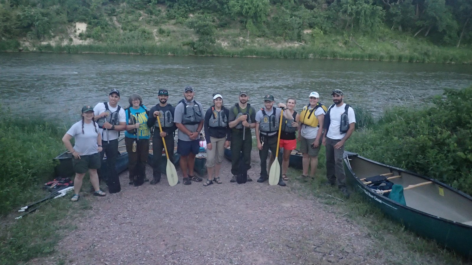 A line of floaters posing for a picture with lifejackets and paddles by a river.