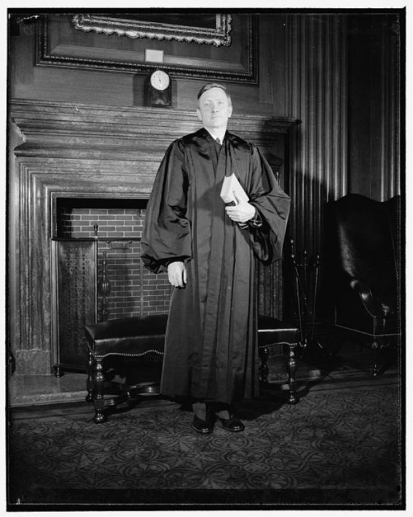 Black and white image of a man in a long, black robe holding the U.S. Constitution