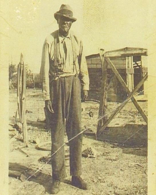 Elderly black man stands in farmyard wearing suspenders and dark pants and light longsleeved shirt