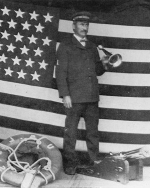 Man in uniform poses in front of US flag with Lyle gun, breeches, buoy, and speaking trumpet.
