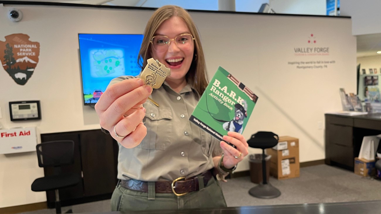 A smiling park ranger holds out a B.A.R.K. Ranger dog tag and activity book