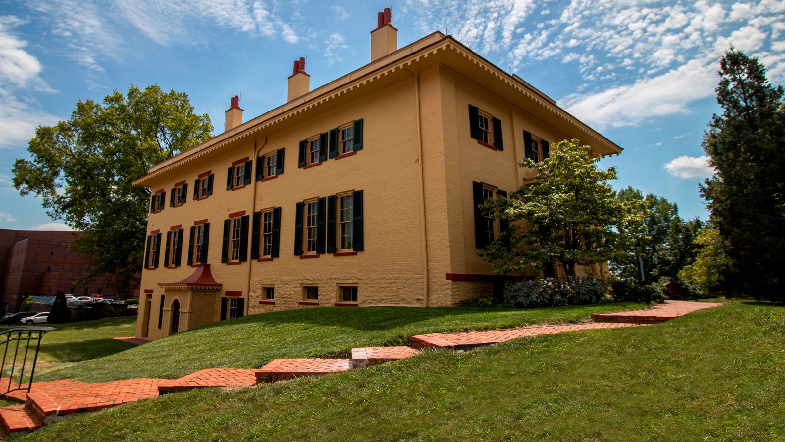 A large yellow house with green shutters surrounding the windows and a brick sidewalk in front