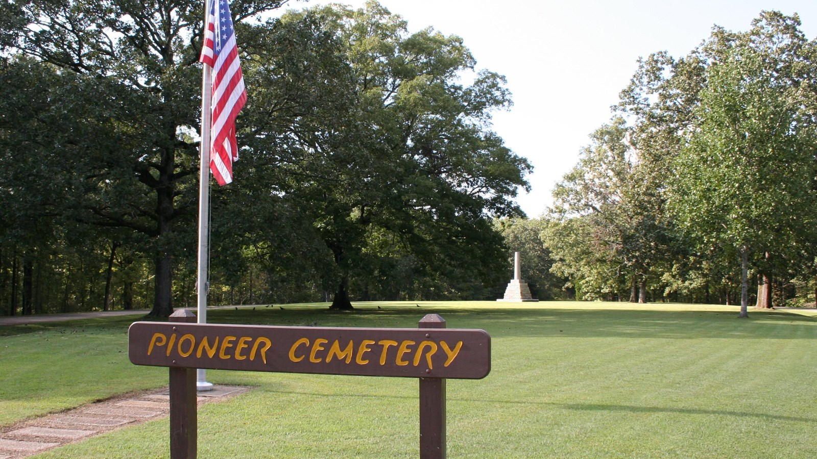 wooden sign painted brown with yellow letters stating Pioneer Cemetery with the cemetery behind sign