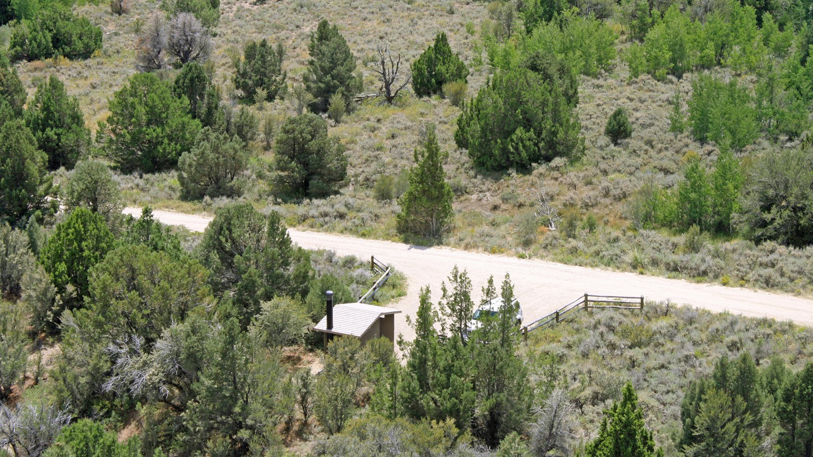 Overview of a vault toilet building nestled among juniper trees and sagebrush. 