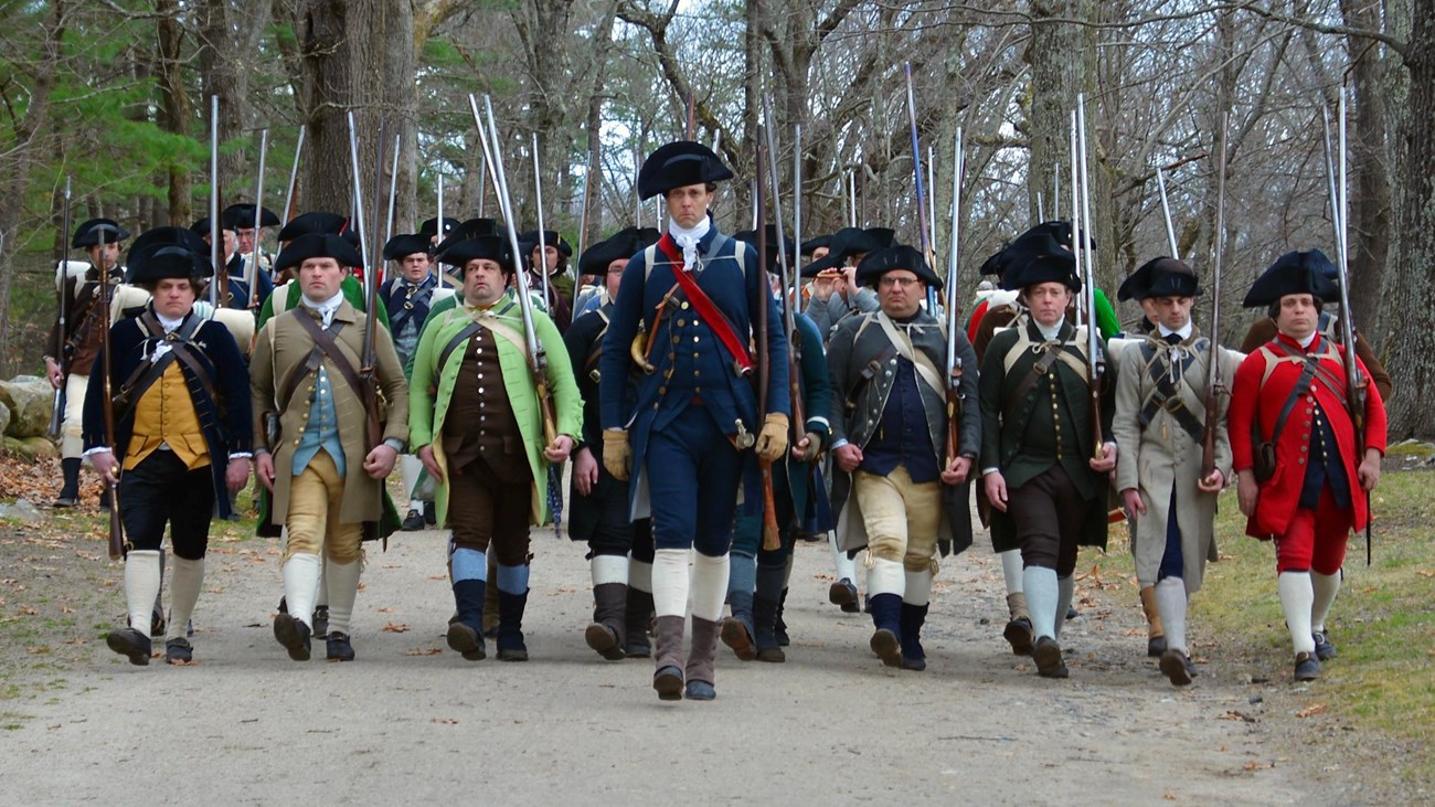 A line of men in 1770\'s clothing march while holding muskets in their left hand.