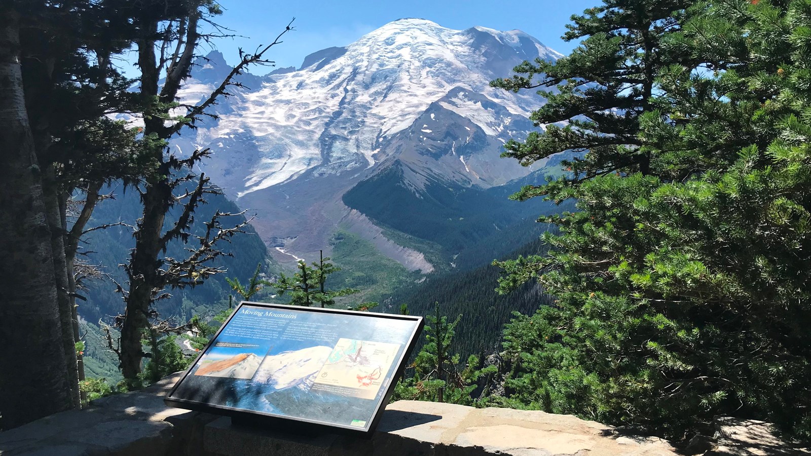 A wayside panel attached to a low rock wall overlooks a deep glacier valley and glaciated mountain.