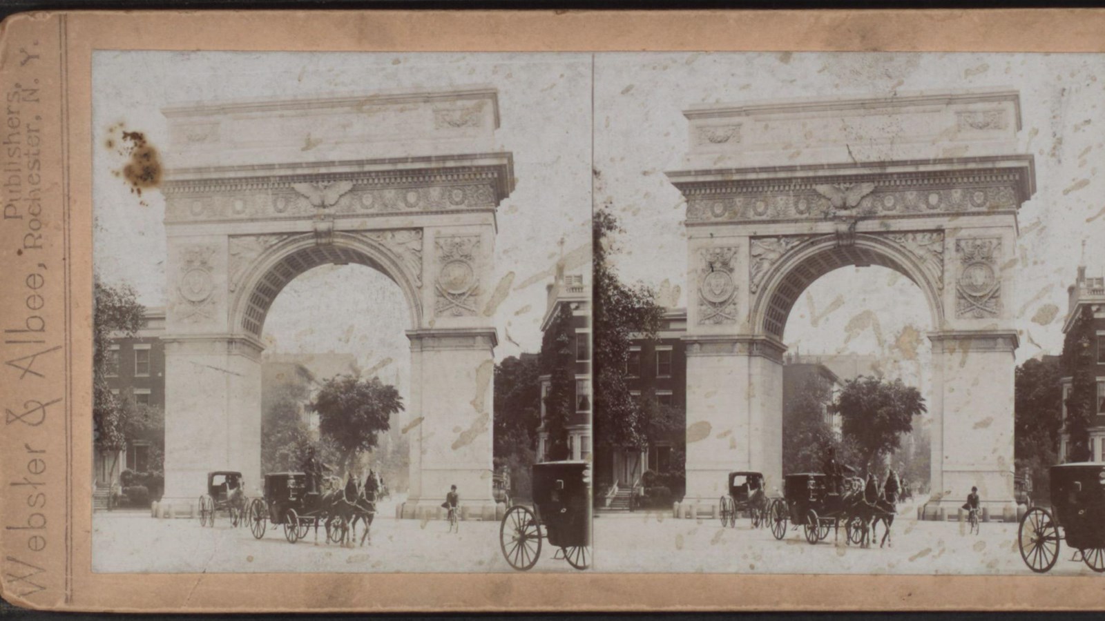 Large decorative arch with horses and buggies passing underneath and buildings in background.