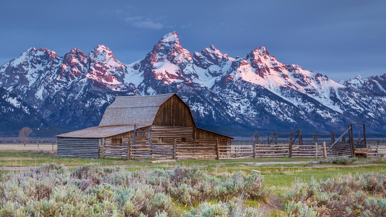 A historic log barn with snow covered mountains in the background.