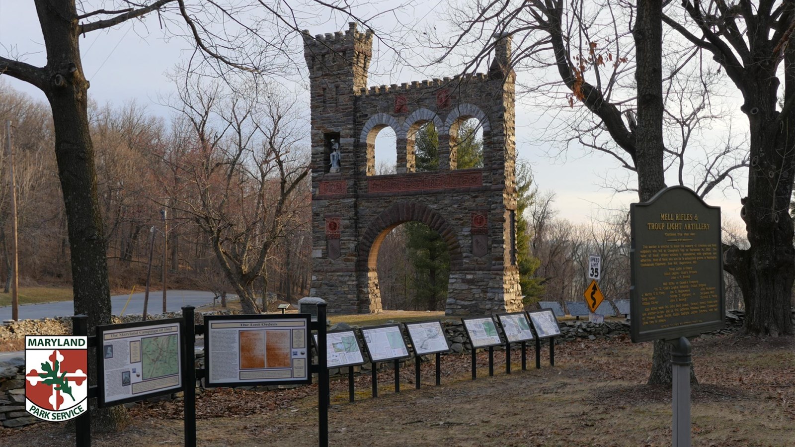 Stone historical arch and roadside interpretive exhibit surrounded by leafless trees.