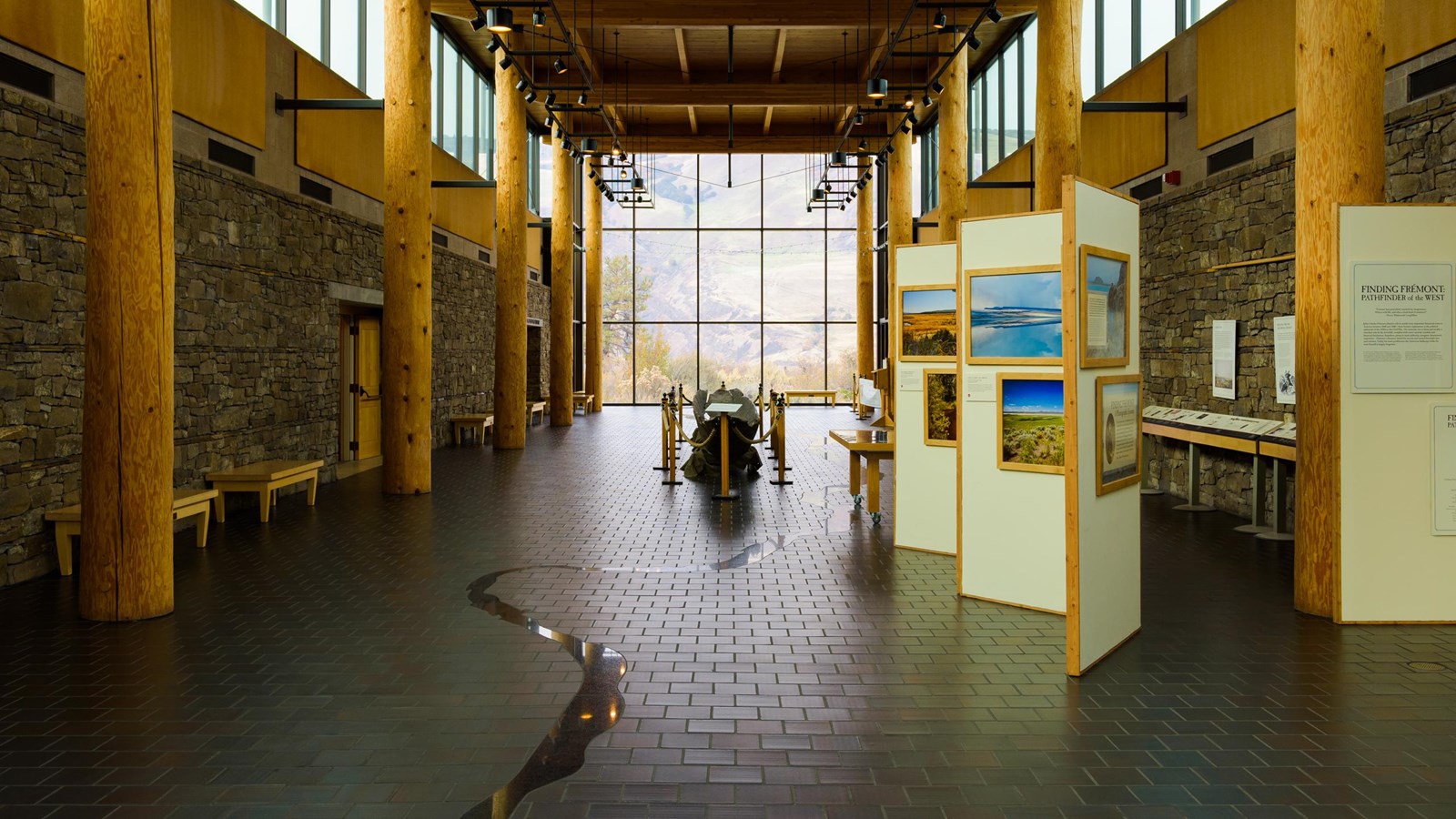 Inside a large room with dark floors and bright windows, upright panels display framed artwork. 