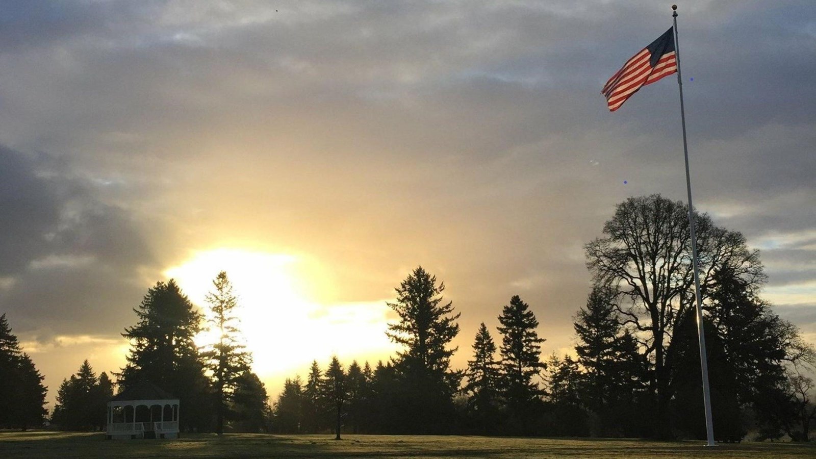 A modern photo shows a grassy field. A tall metal flagstaff with an American flag sits on the right.