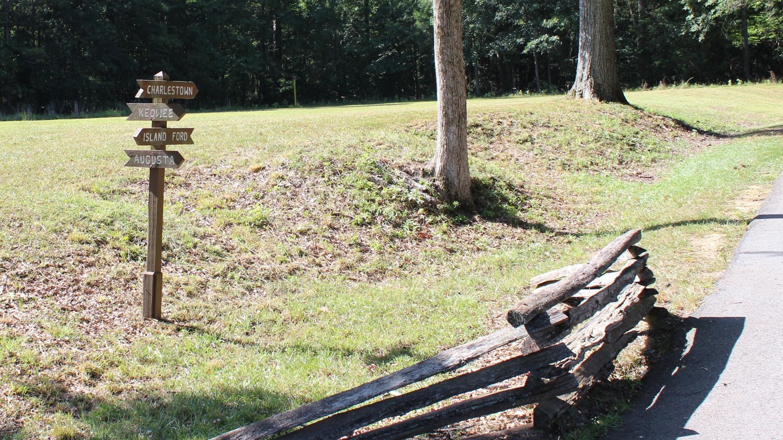 A paved trail with a wooden fence along it with a wood sign pointing in 4 different directions. 