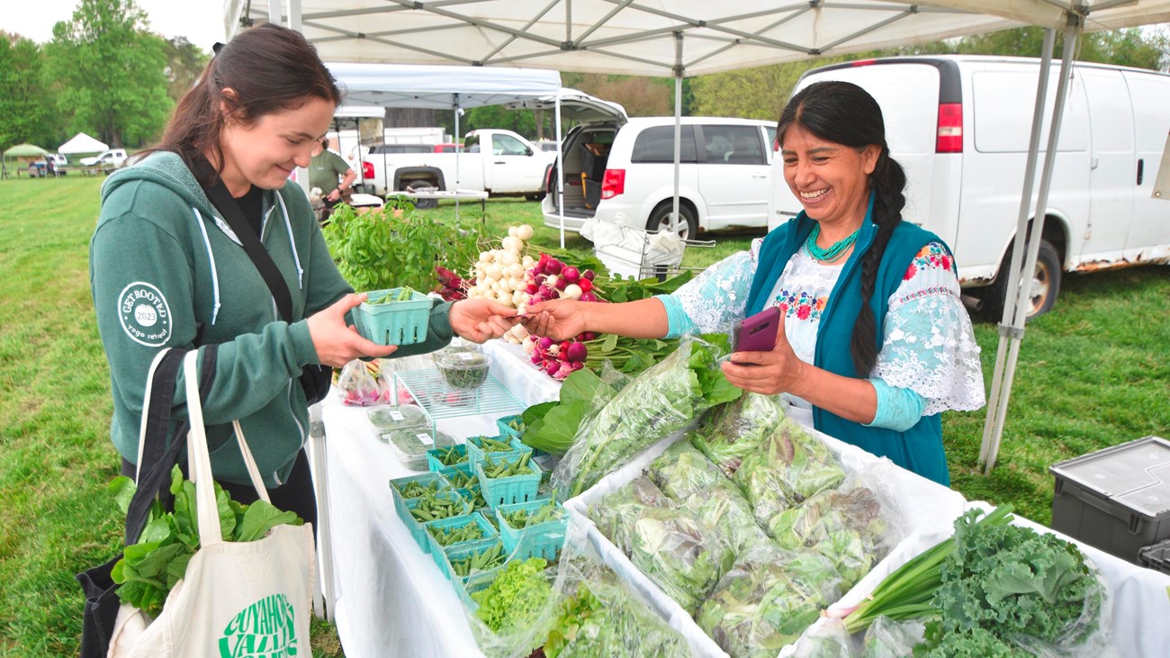 A shopper reaches over table of vegetables to pay a smiling female vendor in embroidered shirt.