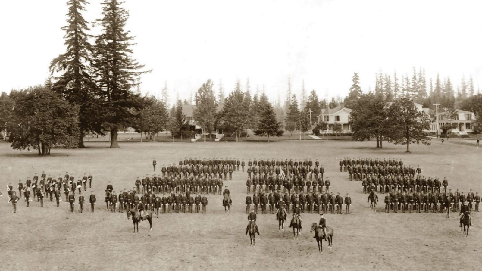 A black and white photo shows soldiers standing in three columns inspected by officers on horseback.