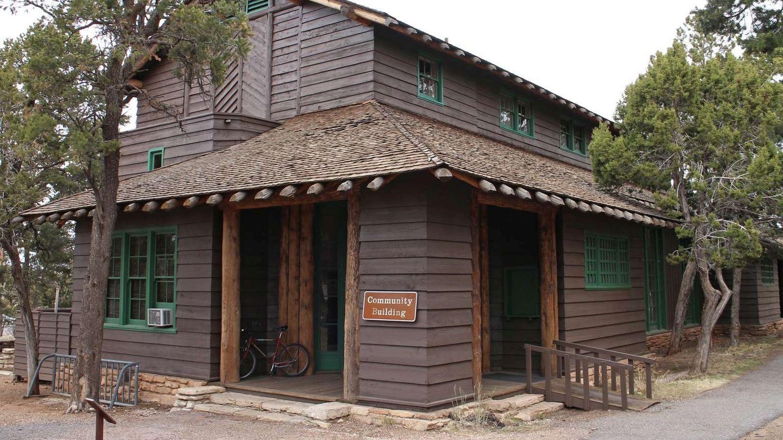 A large brown two story wooden beam building with a shingle room rests among small pine trees.