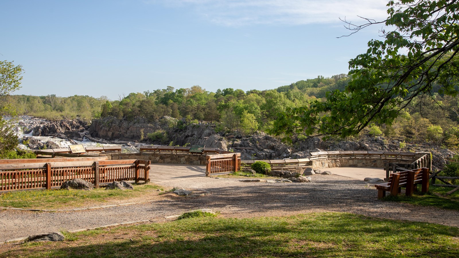 A gravel path leads to a stone and wooden viewing platform overlooking a river lined with trees.