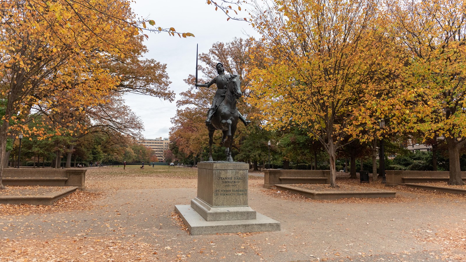 A large statue of Joan of Arc in a plaza surrounded by trees with orange leaves. 