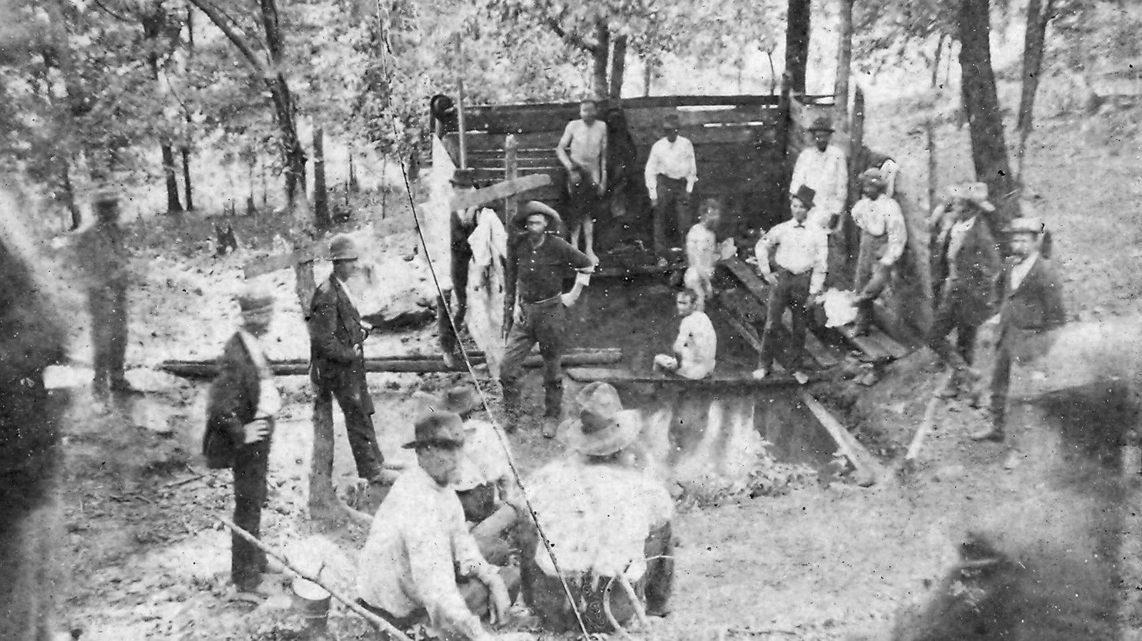 A group of men stand around an open pool of water while one man baths in the pool.