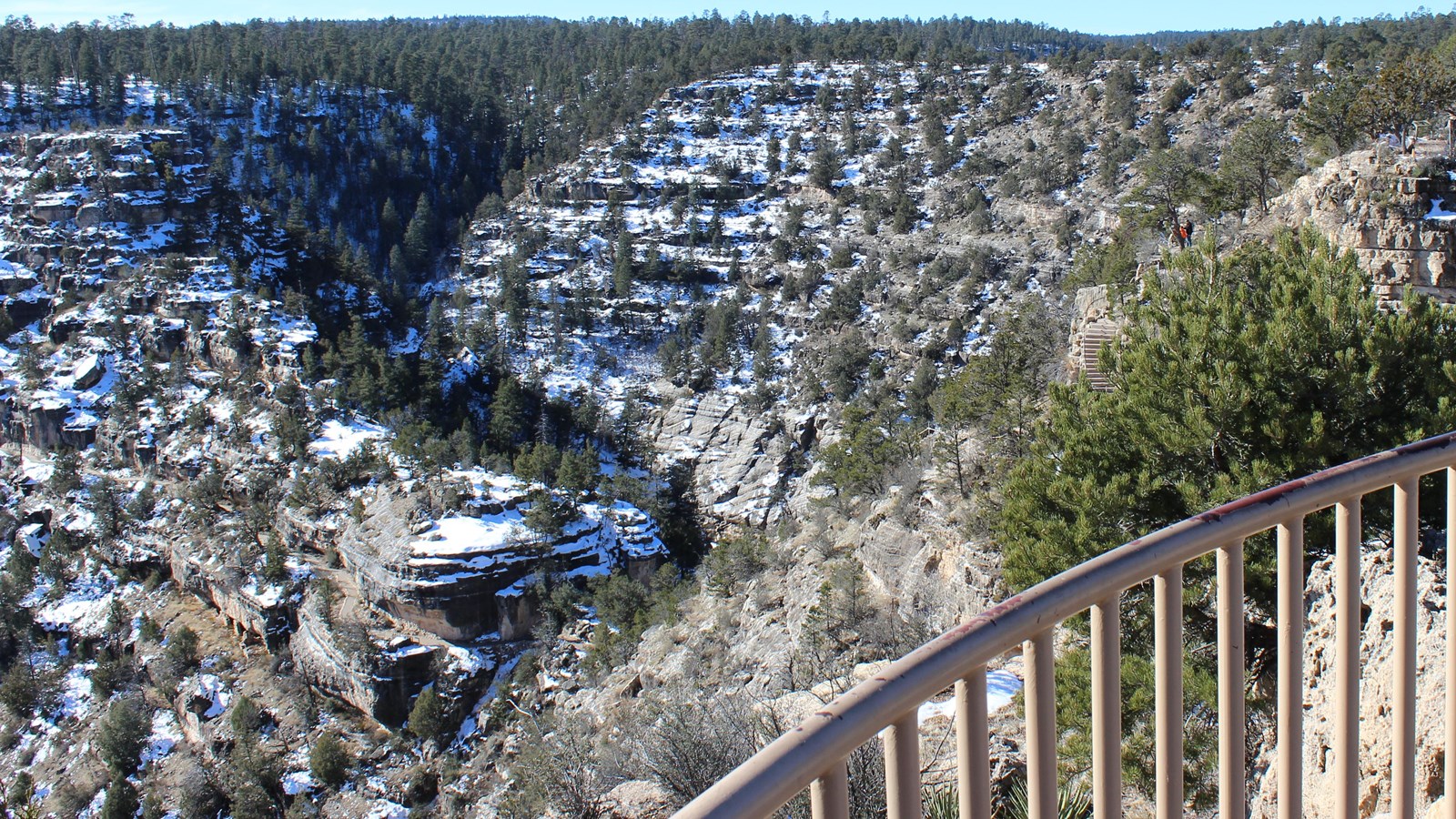 A beige railing overlooking a winding canyon, dotted with green foliage and white snow pockets 