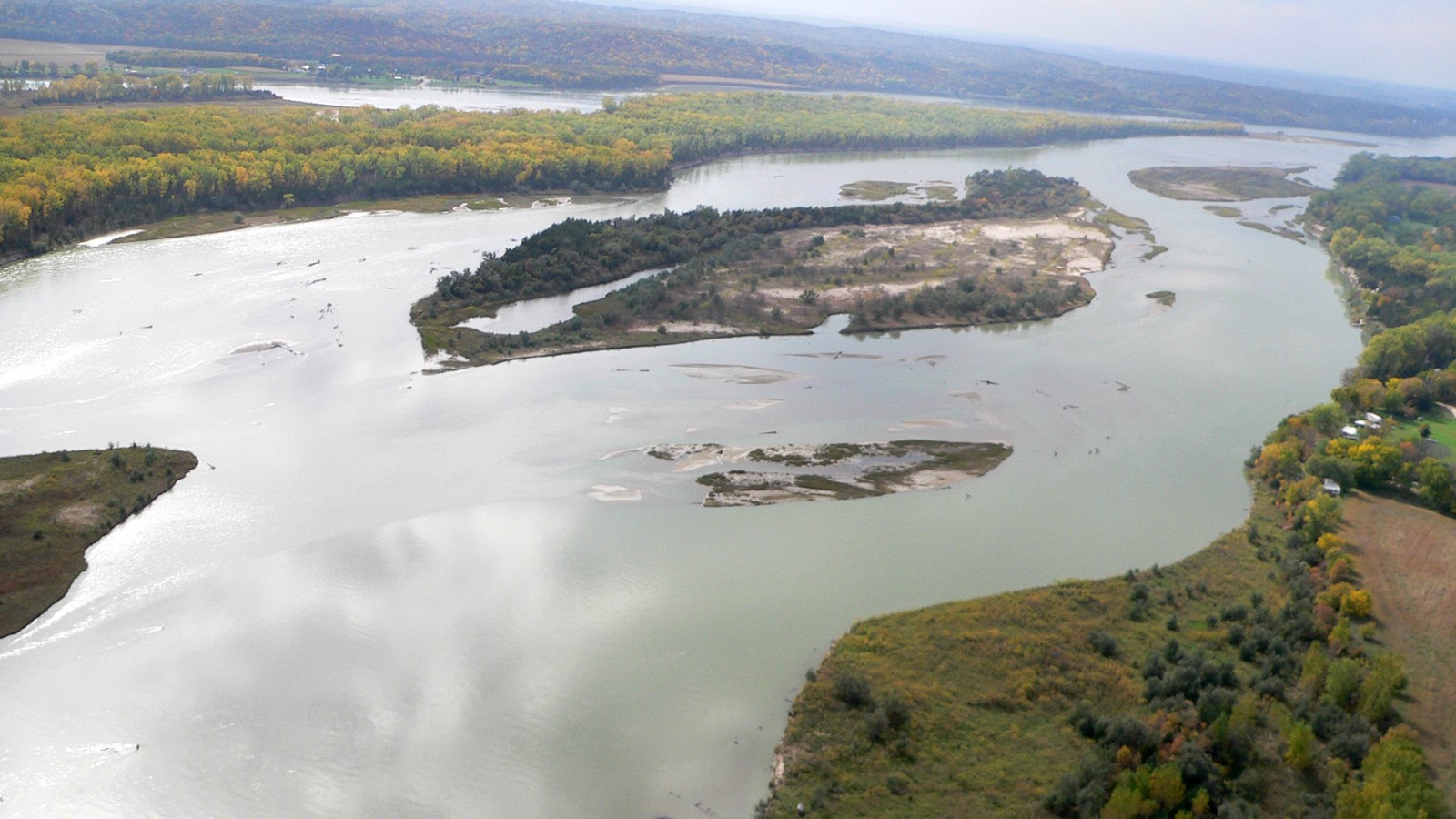 Aerial view of Goat Island with green grass & trees along the bank. River, side channels & sandbars