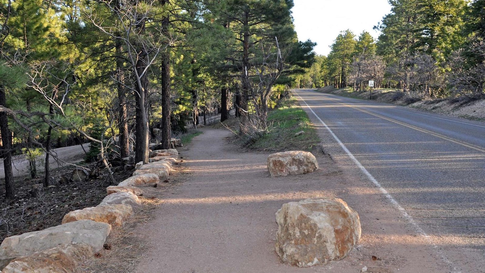 A paved footpath, with rocks lining either side, alongside a 2-lane road in a forested area.