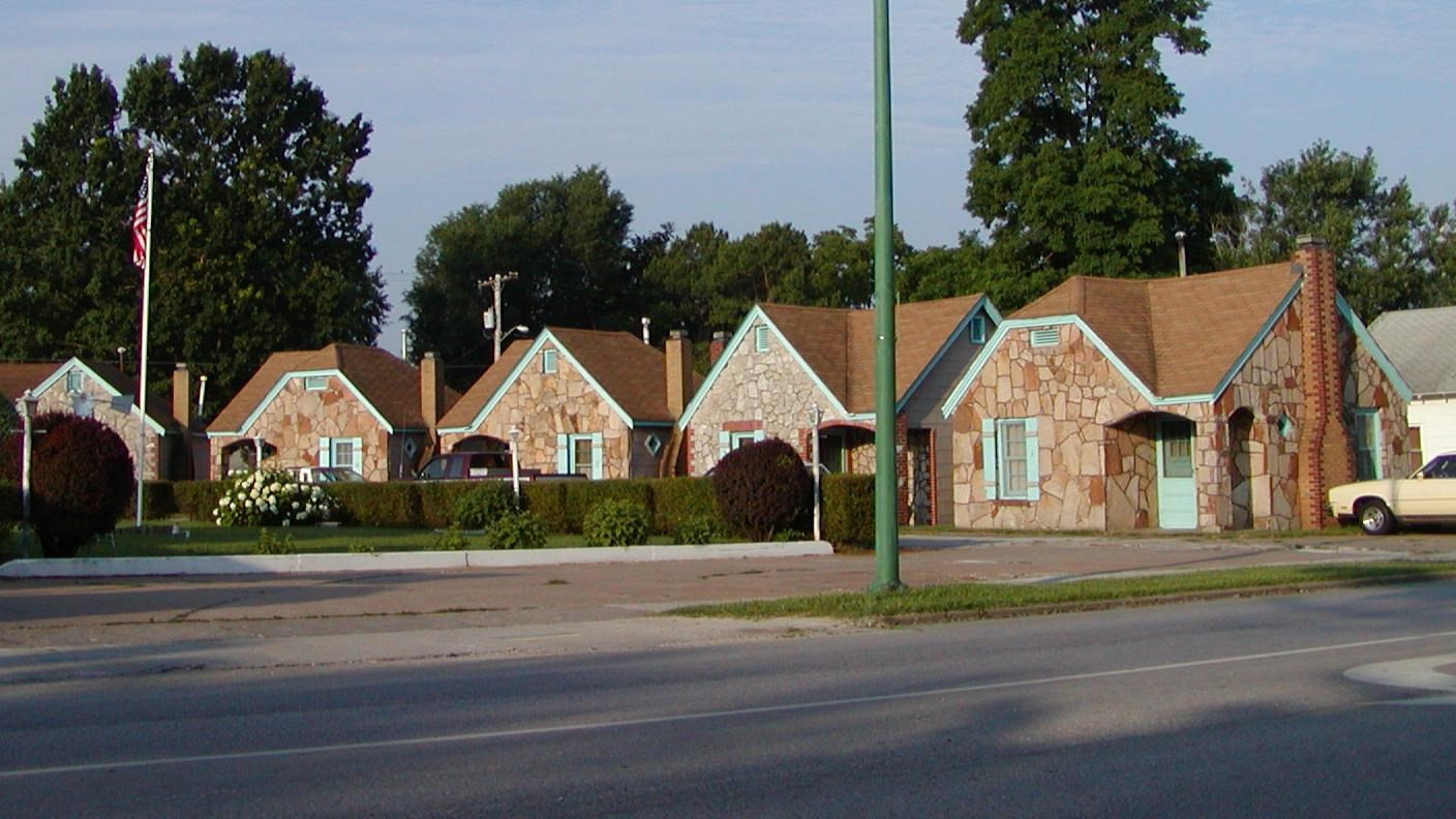 Stone cottages with peaked roofs. Tall trees are behind the cottages.