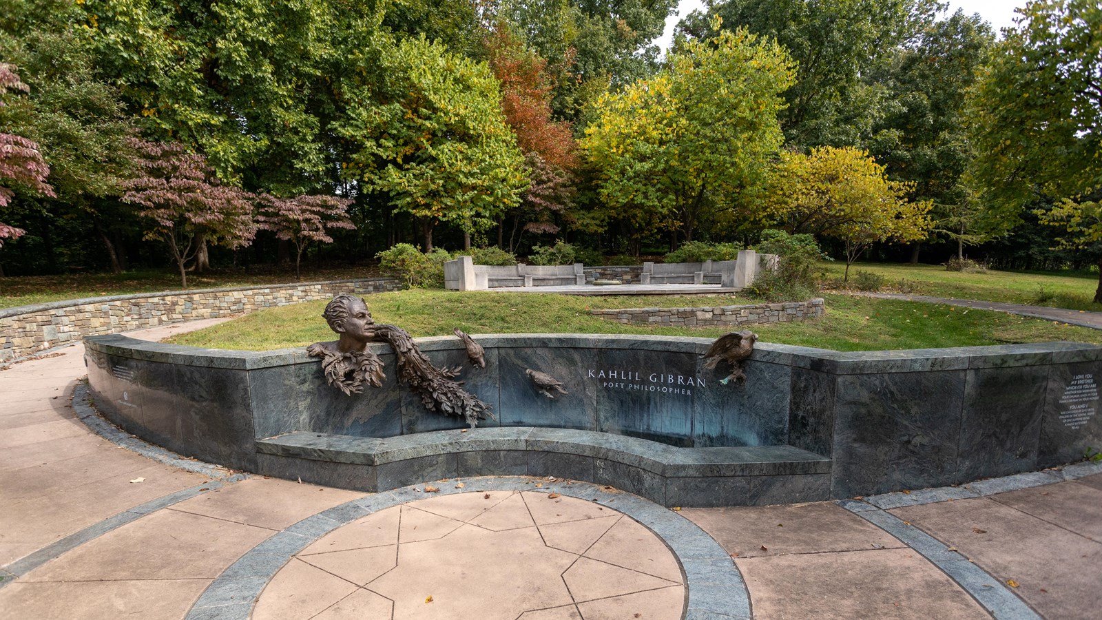 A man on a bench in front of a stone and grassy plaza. 