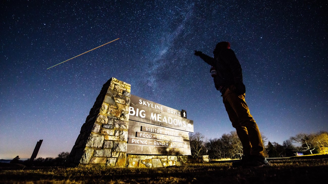 A person points up to the Milky Way Galaxy in the sky over the sign for Big Meadows.