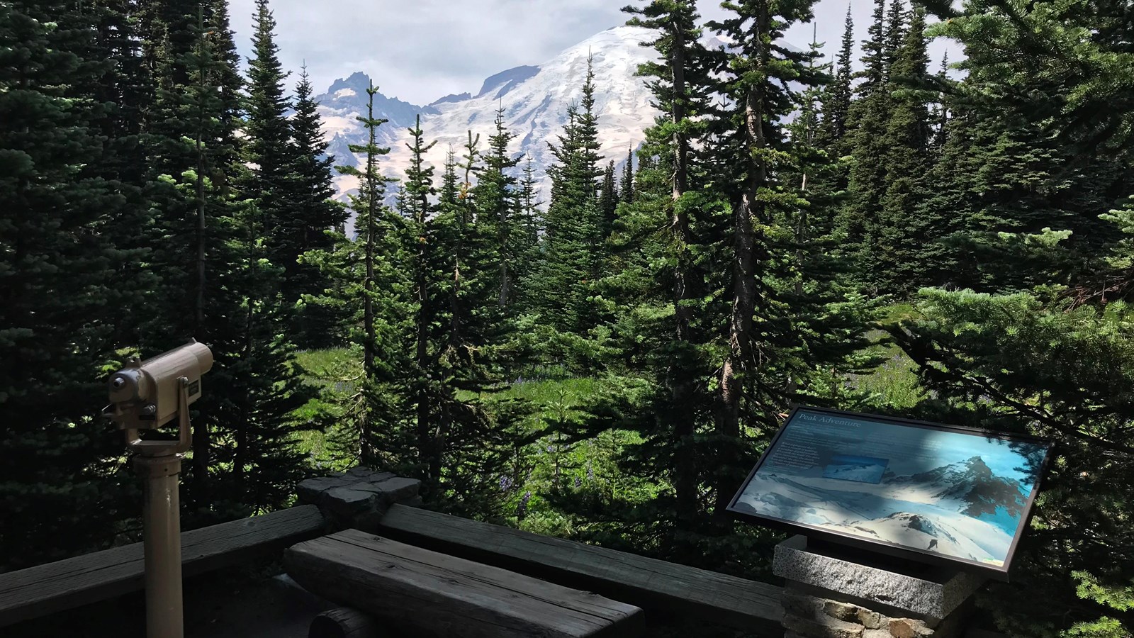 An exhibit panel attached to a low fence next to a spotting scope with a view of Mount Rainier.