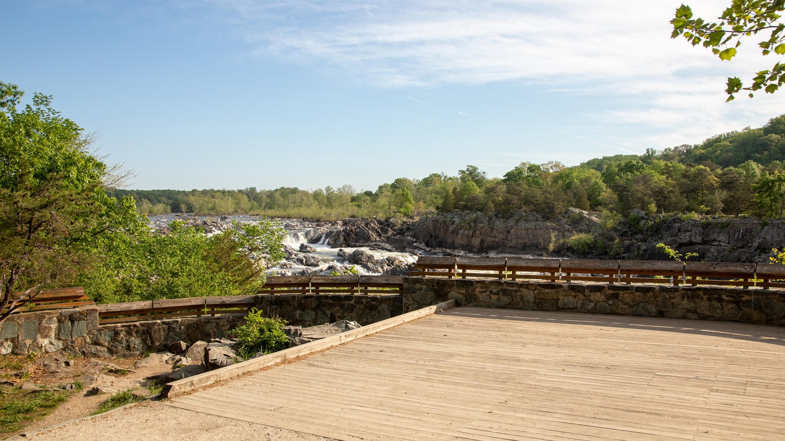 A wooden platform extends out to a stone and wood railing overlooking a rushing rocky river.