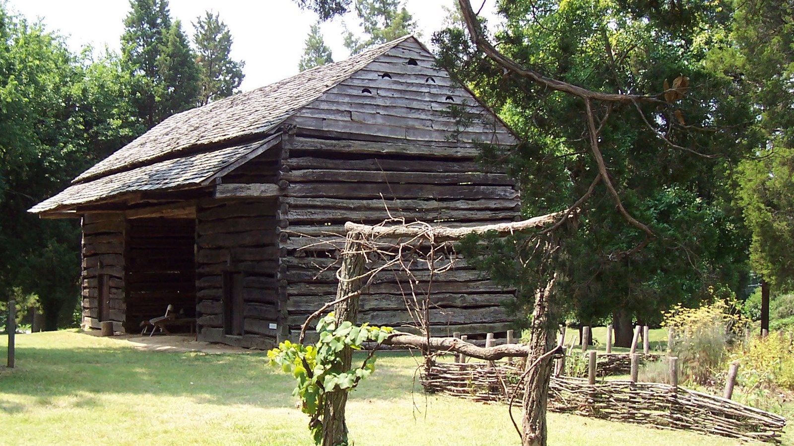 A two story barn with an open log structure. A fenced in garden stand next to it.