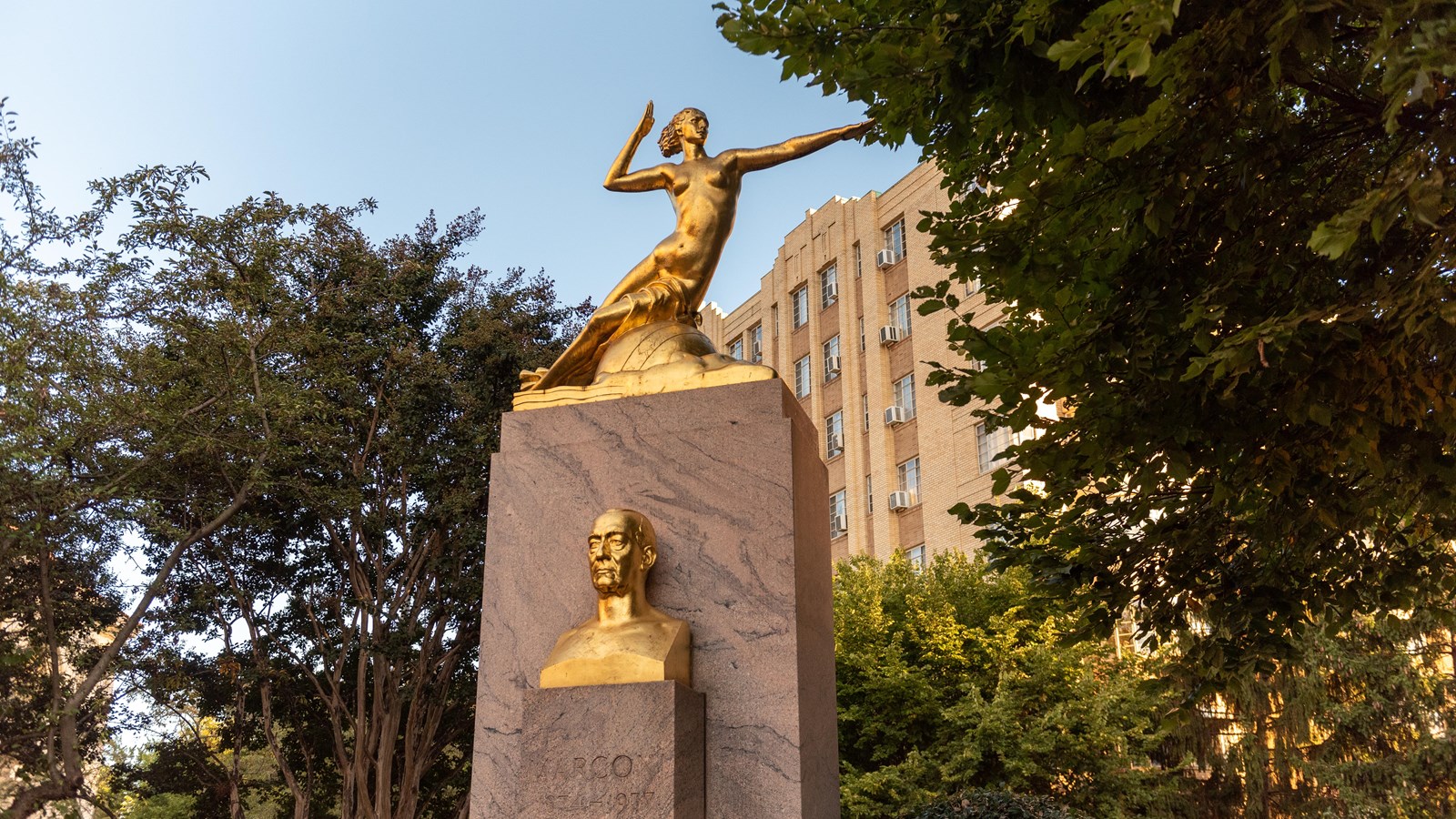 A large golden statue of a woman pointing on top of a golden stone. 