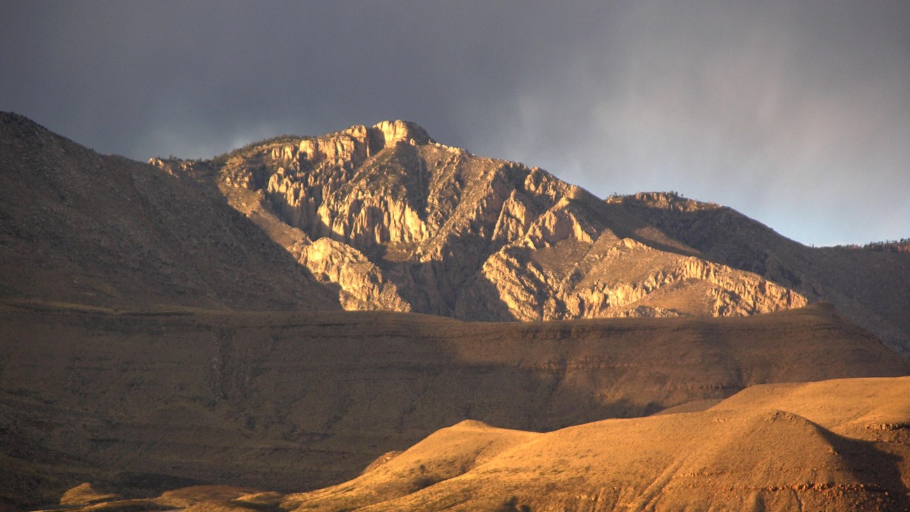 A desert mountain peak is lit by the setting sun as it rises above the landscape around it. 