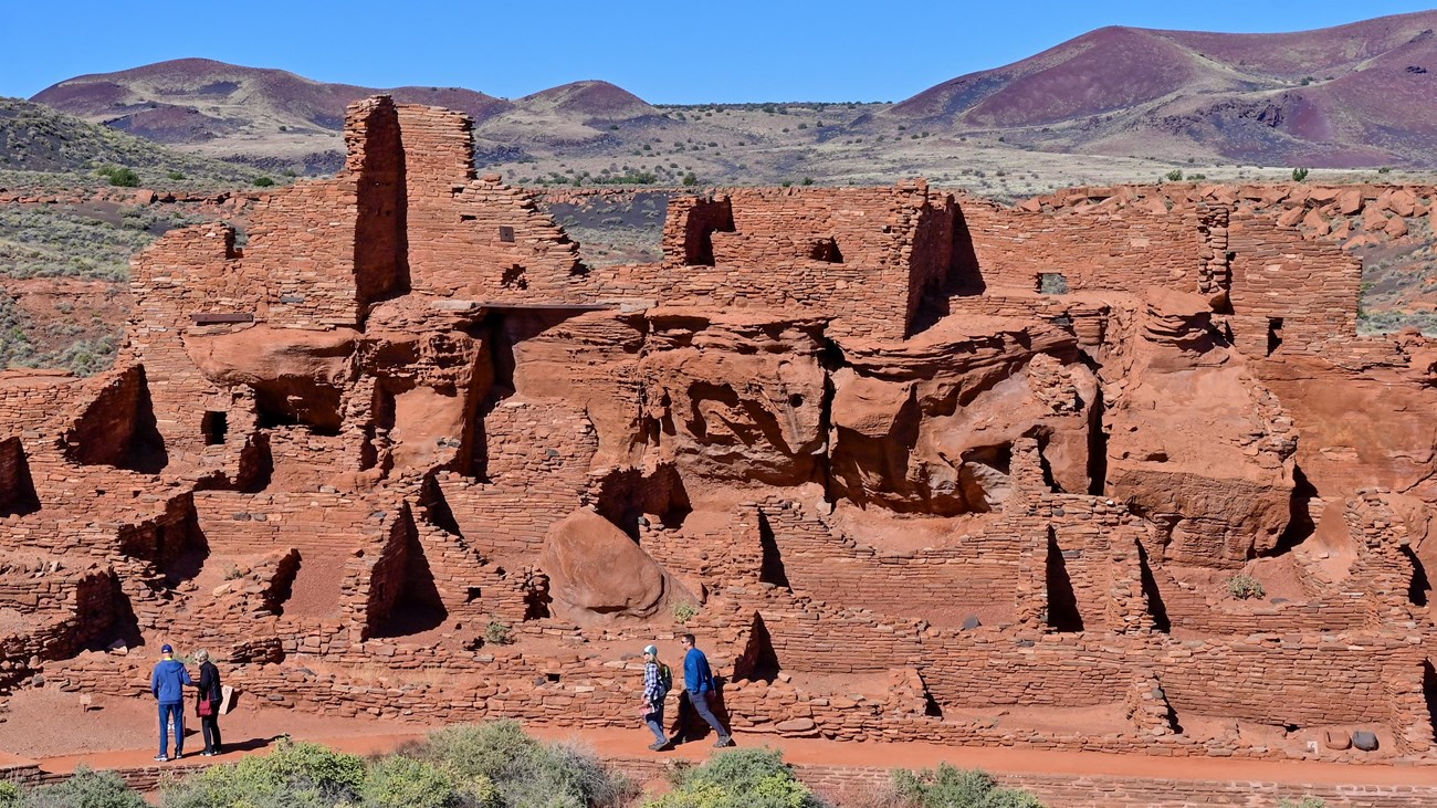 A large multi-room red sandstone pueblo with people walking in front