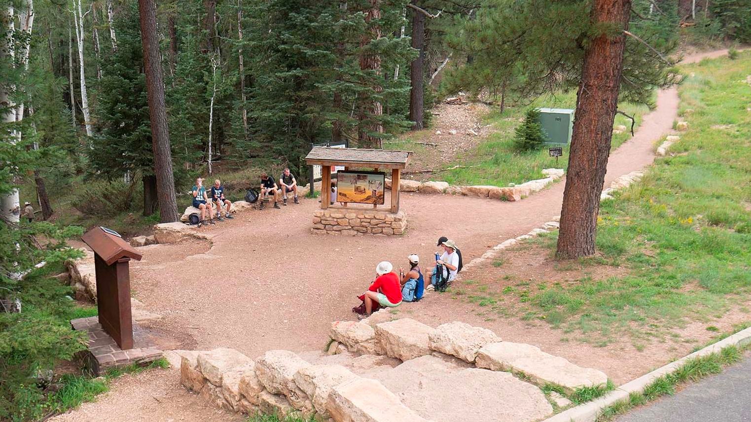 Several people are sitting in a rock-lined open trailhead area, with a footpath on the right.