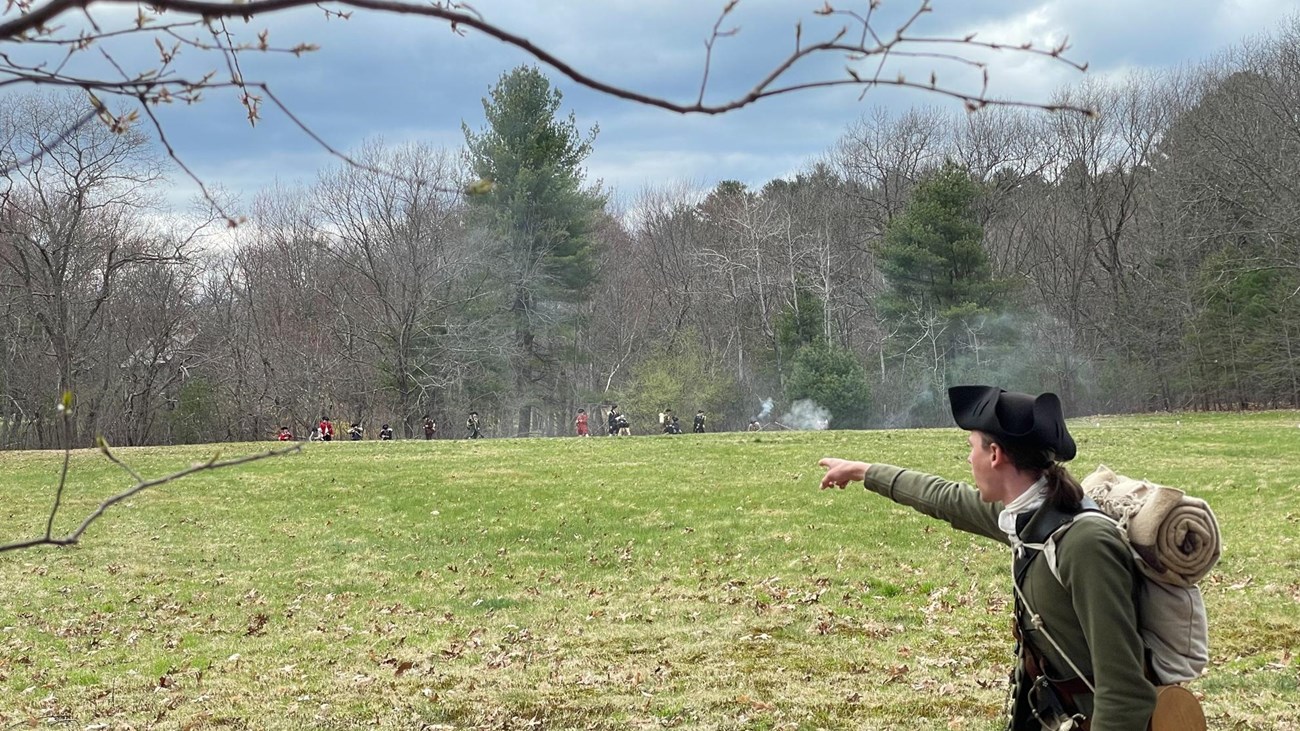 A man in colonial clothing shouts and points across a field at militia soldiers shooting.