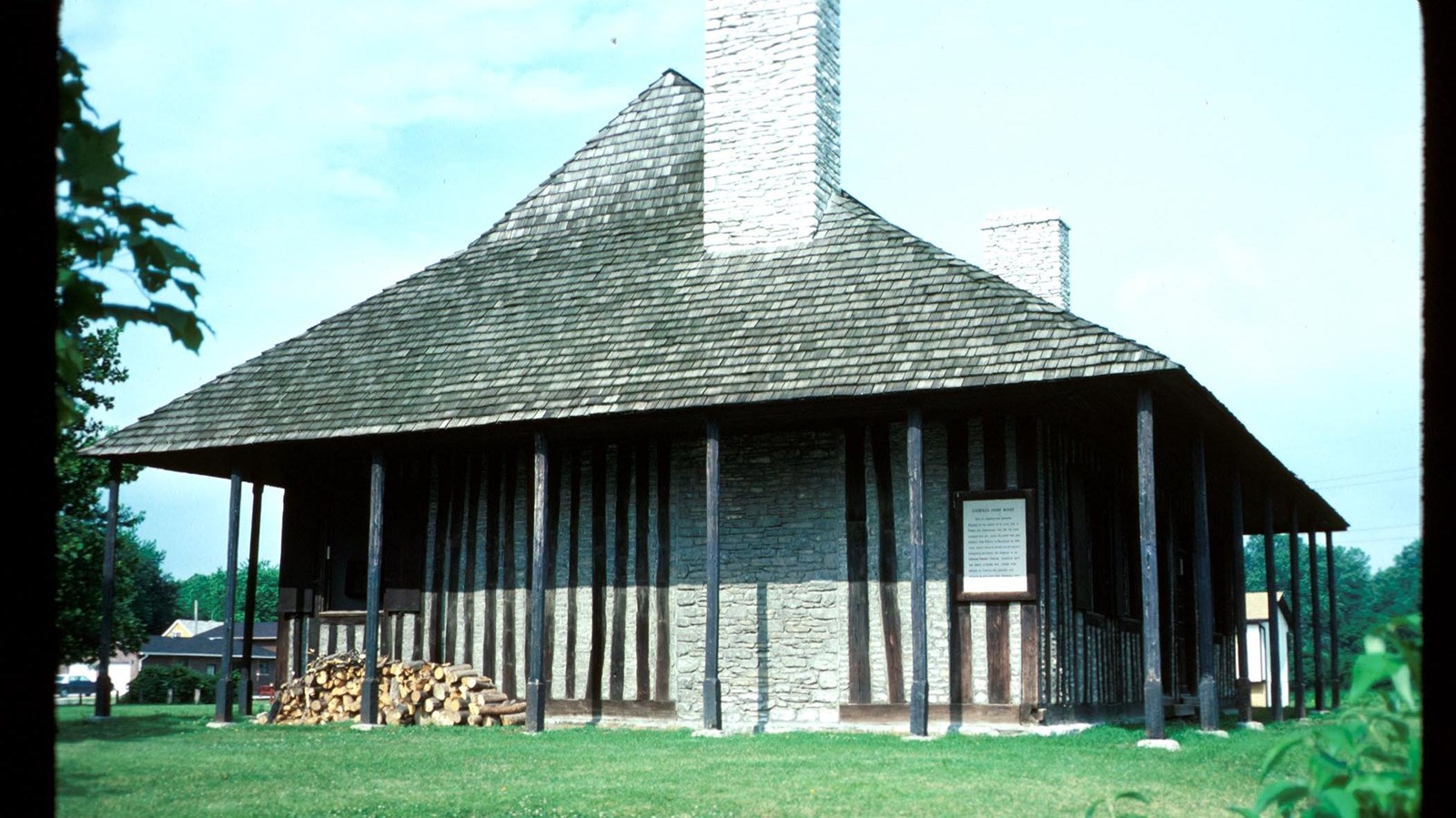 Stone building with wood-shingled roof overhanging a porch and tall stone chimney. 