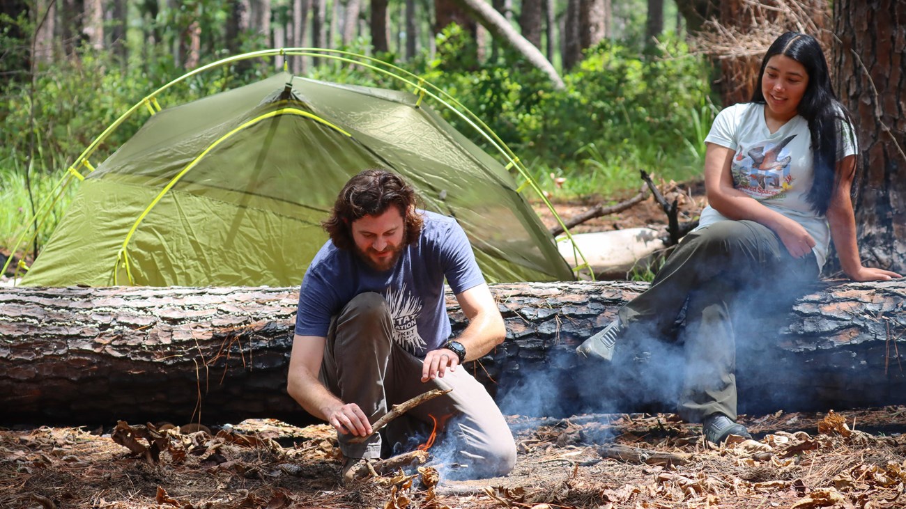 A camper builds a small fire while another camper sits on a fallen log with a tent in background.