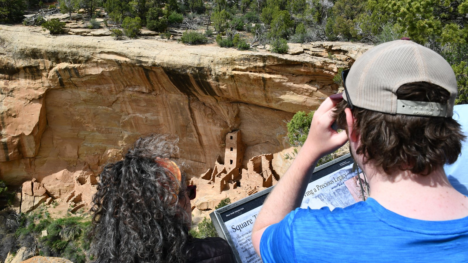 Visitors overlook a four-story stone-built structure beneath a sandstone rock overhang.