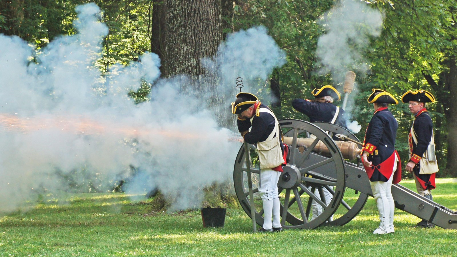 A crew of four blue coated continental reenactors fire a 6 pounder brass cannon.