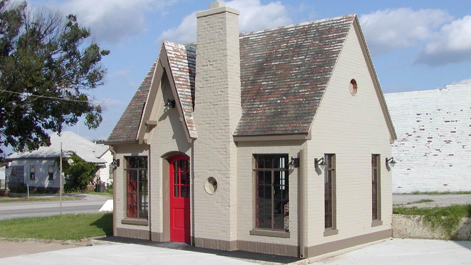 A grey building with a red front door, two windows, a chimney stack and a multi-colored, faded roof.