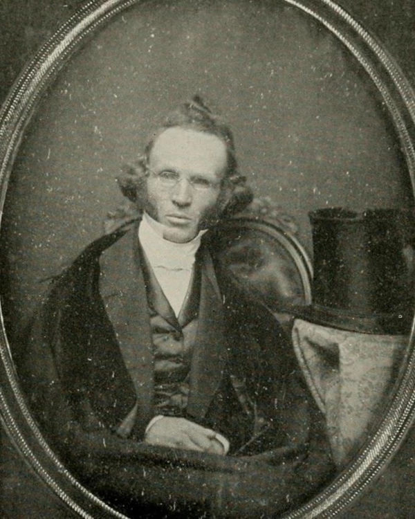 A Black and white photo of a sitting man wearing glasses, with a top hat on a table to his left.