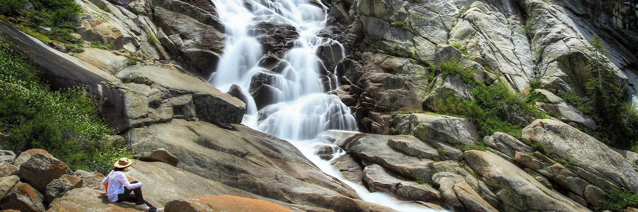 A person sits on rounded rocks at the base of Tokopah Falls.