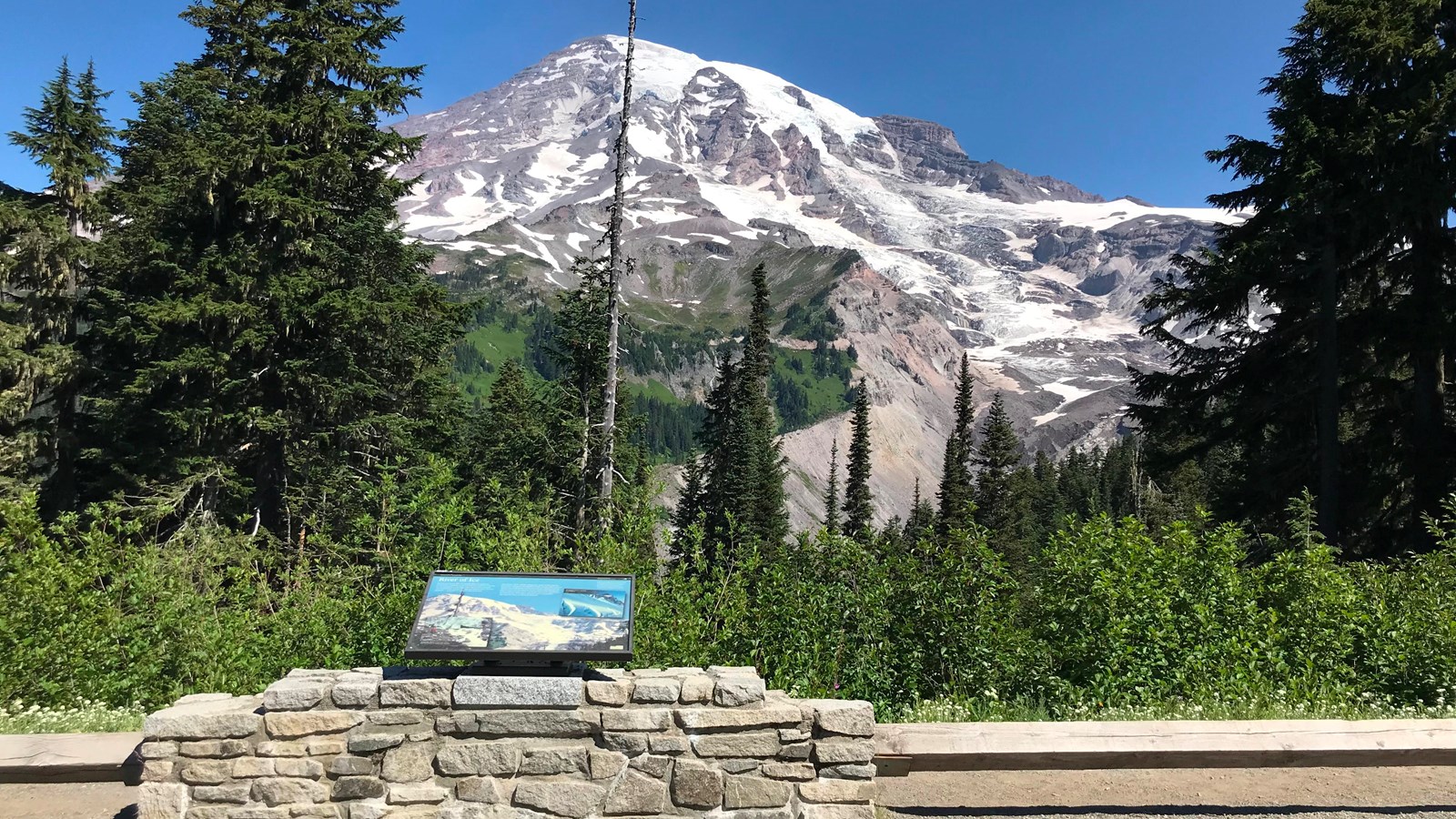 A wayside panel attached to a rock wall in front of a view of Mount Rainier and Nisqually Glacier.