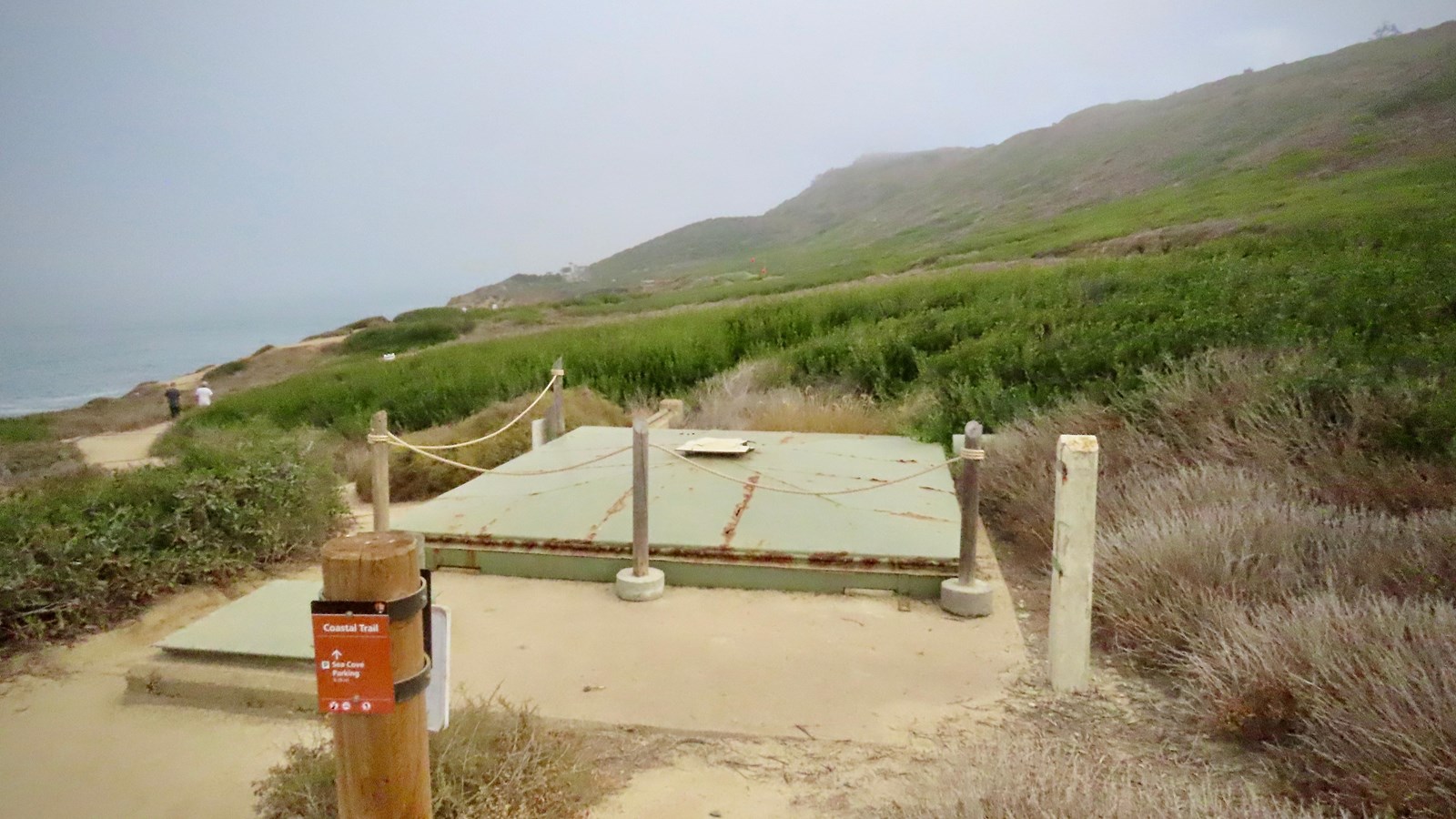 A green metal roof hides a military searchlight along a dirt path. Low green shrubs surround it.