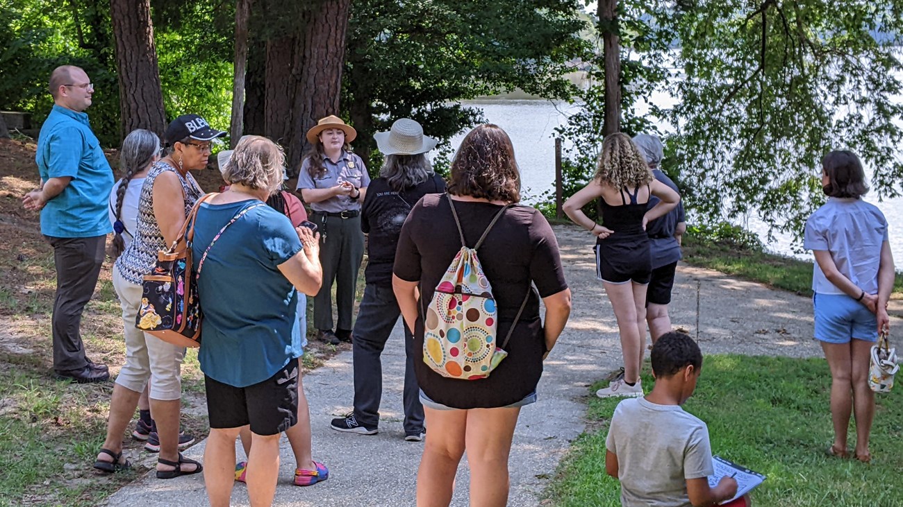 Park ranger standing in front of a group of visitors in the shade with a pond behind them.n
