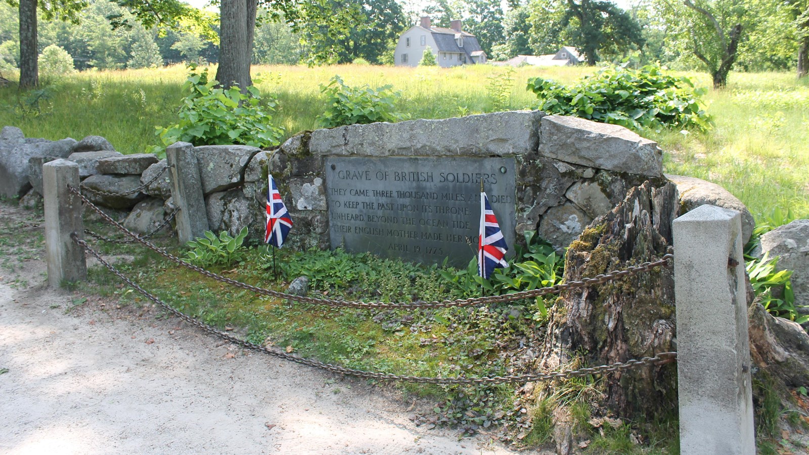 A gravestone is imbedded into a low stonewall surrounded by fencing and green grass.