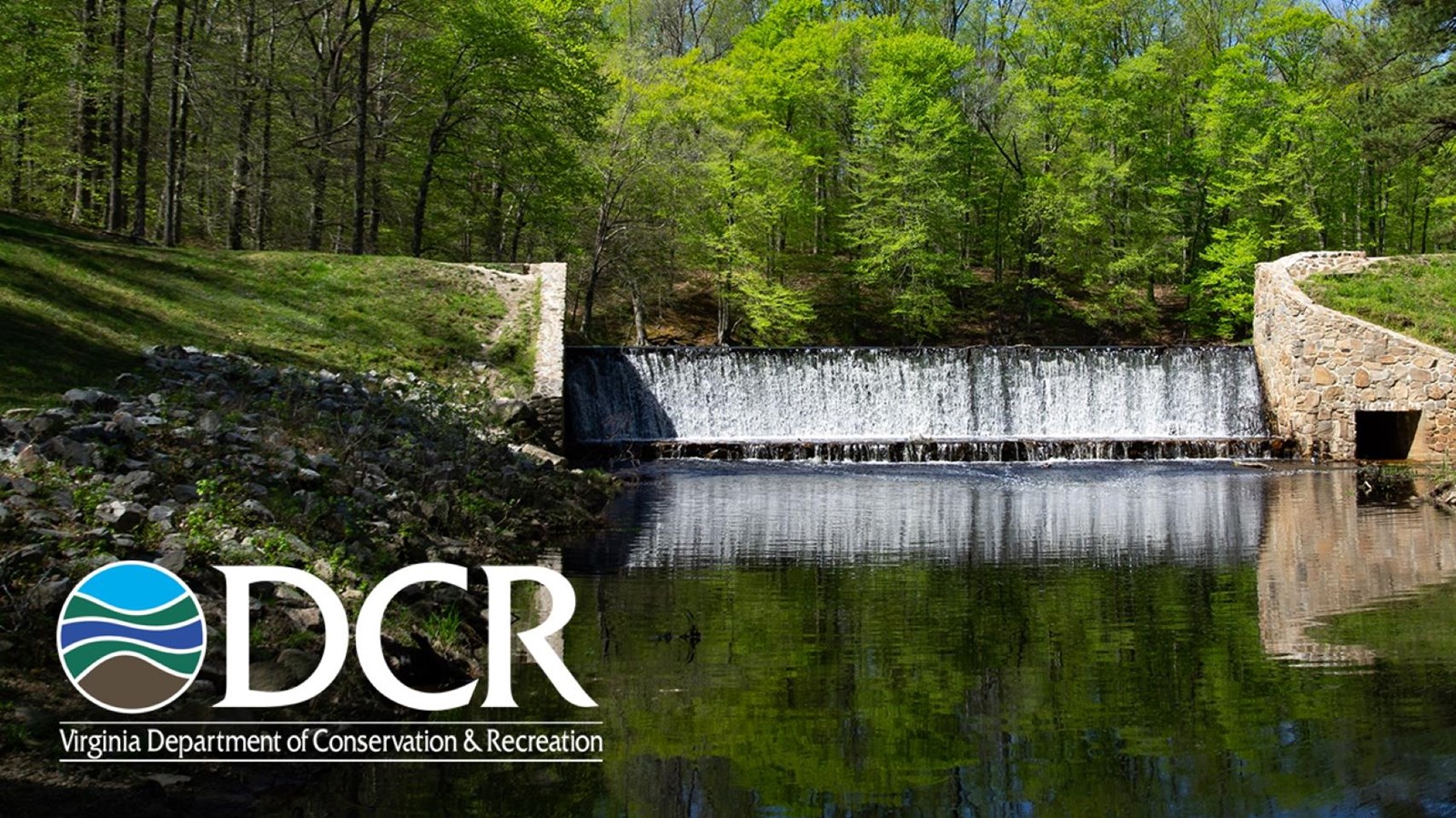 Stone dam and waterfall flowing into a reflective pond in a green forest.