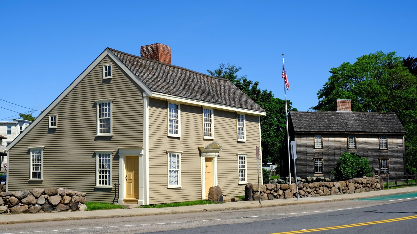 Two saltbox style homes, one gray and one brown, sit next to each other near a road.